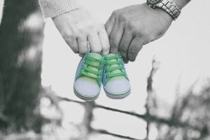 Two hands gently hold a pair of small green shoes, standing out in color against a black and white background with a blurred natural setting. The contrasting colors highlight the tiny shoes, symbolizing the small steps and big milestones to come.