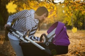 A person tenderly looks at an infant in a purple stroller amidst an autumnal scene with golden leaves. The warm hues of the foliage provide a rich tapestry that signifies the changing season.