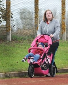 A person pushes a pink baby carriage along a path with a chain-link fence and greenery in the background. The image reflects the quietude of an afternoon walk, the kind that offers a break from the bustle of indoor life.