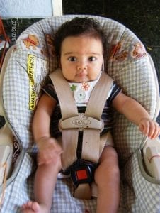 A baby is sitting on a booster chair with safety straps and belt.