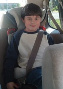 A smiling young boy sitting in the back of a vehicle while wearing a seat belt. Belts are one of the best ways to stay safe during car rides.