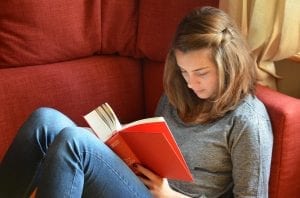 A young lady is sitting on the red sofa in the living room while reading her favorite book. 