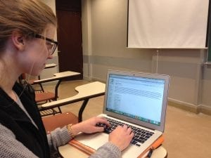 A young woman engrossed in her laptop, fingers dancing on the keyboard. She sits in a cozy spot, surrounded by warm lighting, with a focused expression. The laptop screen illuminates her face, revealing a mix of concentration and excitement.