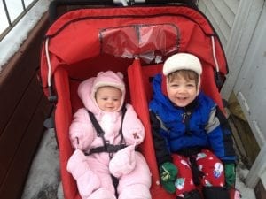 Siblings happily ride the red best double stroller while moving along a narrow pathway of the building. 