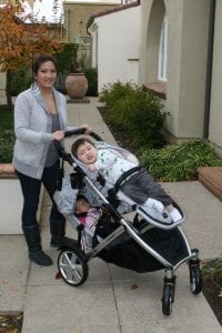 Two kids riding the double stroller comfortably while their mom wearing a gray blazer gently pushes the stroller along the pathway. 