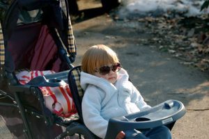 A young child wearing sunglasses sits relaxed in a reclined carriage, bundled up in a white coat, with a clear blue sky overhead and remnants of snow on the ground nearby, suggesting a bright winter day. The child's poised demeanor gives off a vibe of cool confidence, while the carriage stands ready for an outdoor adventure.