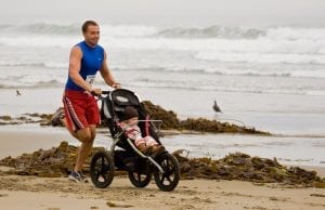 A father with blue shirt and red short joyfully jogs with his baby in a stroller along the sandy beach, enjoying a new stroller with a serene seaside view.