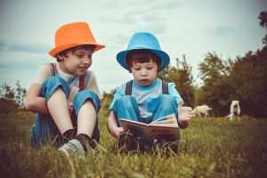 Two boys in colorful hats reading one of the best books for their age, sitting on a grassy field under a clear sky.