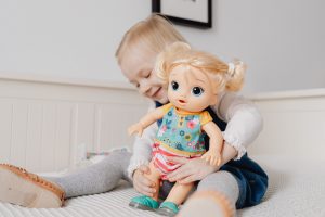 A kid happily playing with her favorite baby doll inside the bedroom. This baby doll can serve as a comfort object, offering a sense of security and familiarity, especially during times of transition or new experiences. Additionally, it becomes a cherished companion fostering emotional development. Furthermore, engaging with a baby doll in play contributes to the development of essential life skills, such as empathy, responsibility, and nurturing, laying a foundation for positive social interactions in the future.