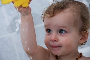 A toddler is enjoying her bath. You can see that she has red spots on her face because of sensitive skin. Using a nice brand of lotion for this can be a big help.