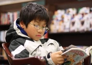 A boy who is setting on a chair who is seriously reading a literary piece intended for male student like him in a quiet library