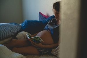 Inside a dark room, a pregnant mom is reading a literature about pregnancy leaning on the wall while setting at the bed with many pillows.