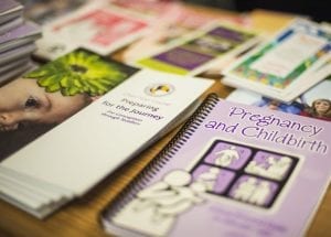 A spring notebook with a label "Pregnancy and Childbirth" and different flyers are on the brown table. All literature are for expectant moms.