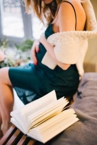 A brown-haired mom on her sexy green dress is setting on the bed with a book open.