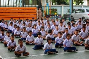 Children wearing school uniforms sit on a cemented ground. 