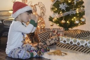 A 4 year old boy wearing pajamas and a Santa hat is opening the presents he has received for Christmas. The child is also drinking milk on his sippy cup.