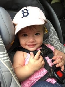 A baby with a white hat is seating in a car with a smiling face in her car seat with a 5-point safety harness-the safest for kids age 3.