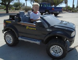 Power Wheels: A young child sits in a black Power Wheels ride-on toy modeled after a Ford pickup truck, with the name "Gregory" on the side.