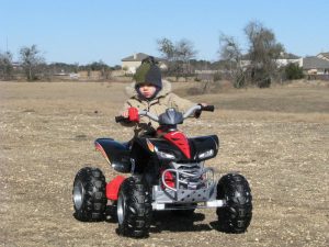 A young child, bundled up in winter clothing, rides a red and black Power Wheels ATV in an open field.