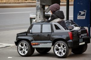 Power Wheels: A toy Power Wheels SUV with a flame design is on the sidewalk, beside an adult at a utility pole.