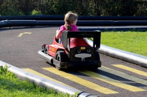 A child drives a red and black Power Wheels racing car on a track with lane markings.