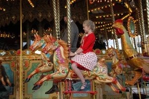 A 9-year-old girl with a bright red shirt sits on a colorful carousel ride at an amusement park. The carousel features intricately designed horses and other whimsical creatures, each painted with vibrant colors. The girl wears a delighted expression on her face, her hair flowing in the wind as the carousel spins. The background showcases a bustling amusement park, with other rides, people, and joyful atmosphere.
