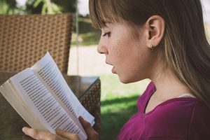 A young girl in a purple top is reading a spanish book for fun, with natural light illuminating the pages.