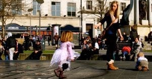 A young girl in a pink dress runs joyfully in an urban square, there are moms and other pedestrians in the background, enjoying a sunny day outdoors.