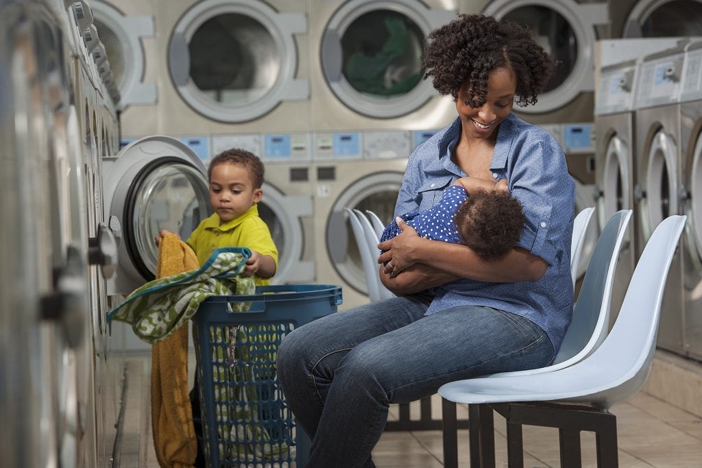 A woman carrying a baby is in the laundry room with another child.