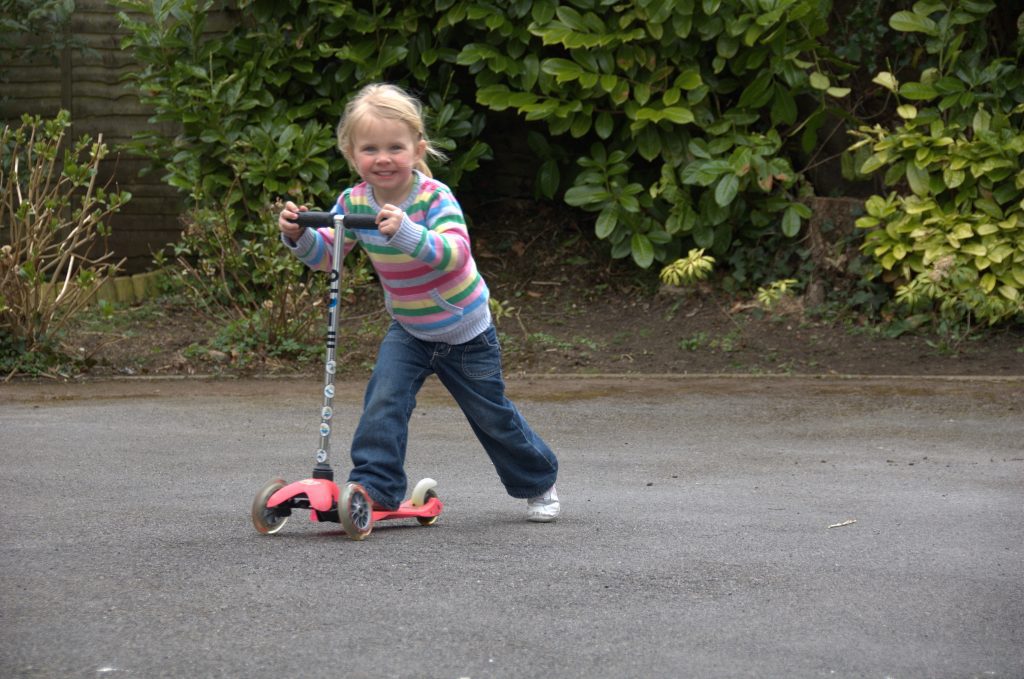 great scooter, toddlers scooter or kids scooter: A child with her scooter, smiling wide while riding in her scooter playfully. 