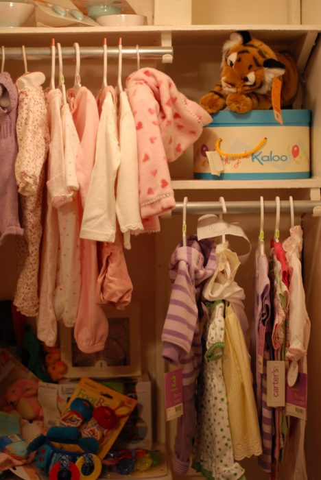 A well arranged cabinet containing different things for infant such as a tiger stuffed toy, a box with yellow handle, pink and white clothes hook with different colorful baby's clothes hanged on it. 