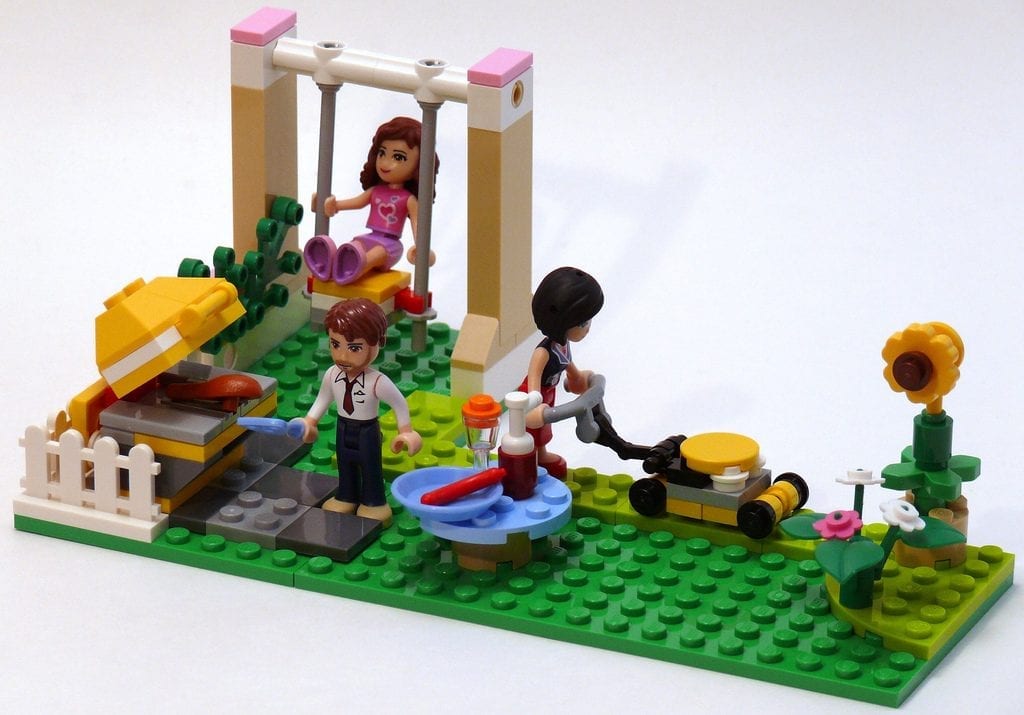 A vibrant LEGO Friends playset depicting a bustling park scene, complete with a detailed swing where a mini-doll figure enjoys the play area. The set also includes an interactive BBQ grill with food elements, a character grilling, and a lawn mower being pushed by another figure. This setup is enhanced by a colorful garden area with flowers, symbolizing a perfect day out in a LEGO town.
