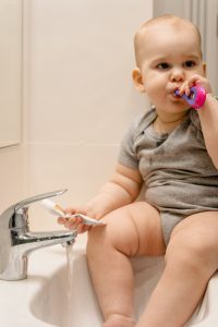 A baby holding his best toothbrush sits on the sink in the bathroom and brushes his teeth with that toothbrush. The toothbrush is colorful making that toothbrush attractive to the baby. 