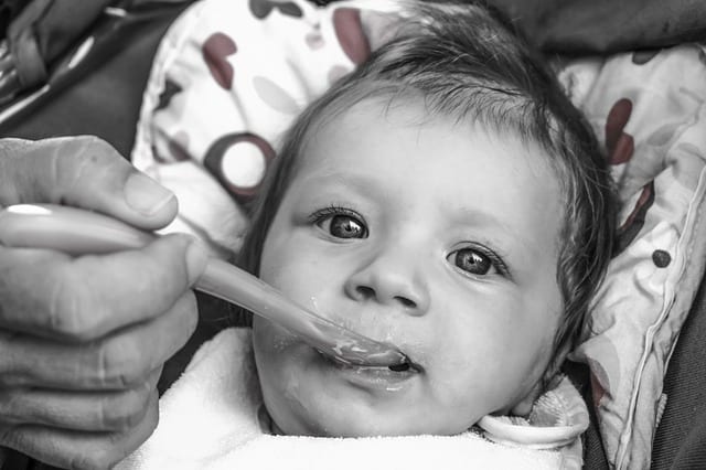 A gray picture of a hand of a mother feeding her baby with round eyes with his favorite food using a baby spoon while looking in front of the camera. Feeding babies with the right baby spoon is essential.