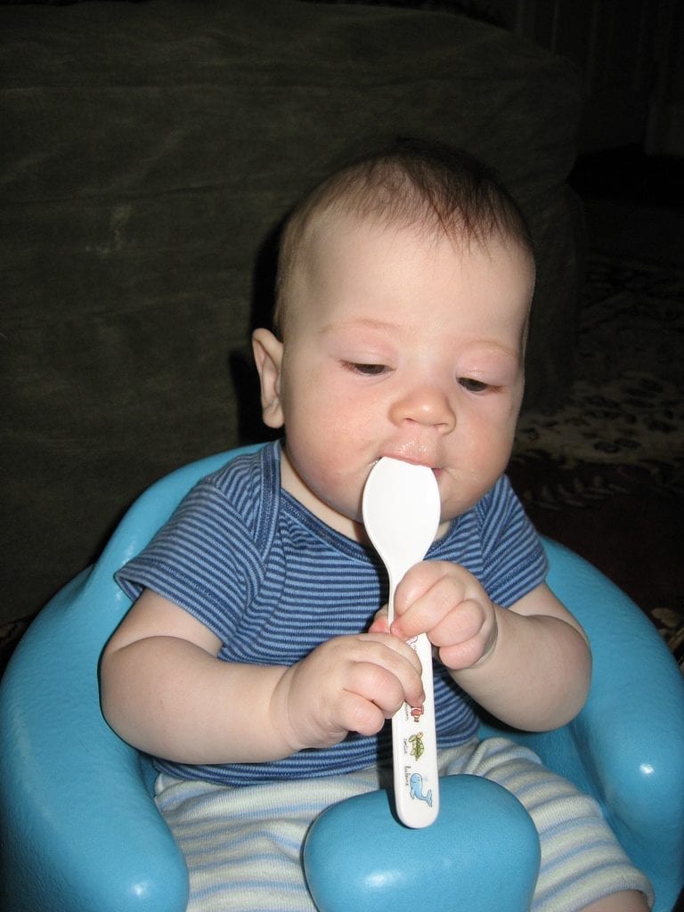 An infant setting on a blue chair wearing blue stripe shirt holding a white best baby spoon and put it on his small mouth while enjoying the moment. This white best baby spoon is just perfect with his cute little hands.