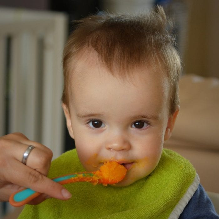 A mom holding a baby spoon pointing it to his baby's mouth letting her baby with brown hair and round eyes eat his favorite food. This best baby spoon with color blue and orange combination looks great with the orange baby food in it.