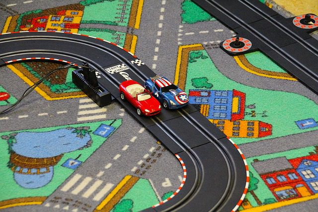 Two car toys arranged on one of the race track toys placed on a carpeted floor inside a bright room.
