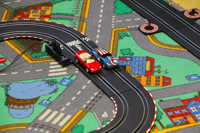 Two car toys arranged on one of the race track toys placed on a carpeted floor inside a bright room. 