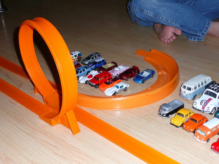 Small toy cars are arranged together on the floor near the orange racing track while a little kid is playing with the toys. 