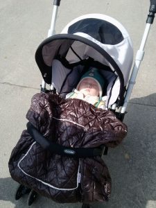 Best Car Seat Stroller Combo: A cozy baby bundled up in a quilted brown bunting bag, securely fastened into one of the best car seat stroller combos, featuring a sleek grey and black design, ready for a safe and comfortable stroll.