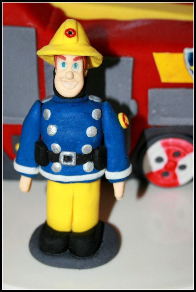 Fireman Sam: Fireman Sam toy figure in blue uniform and yellow helmet, standing in front of a red toy fire engine.