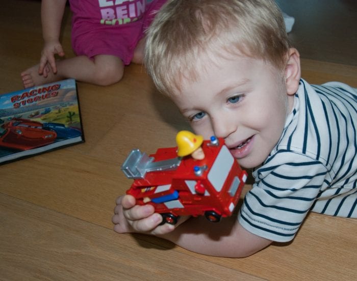 Fireman Sam: A smiling child lying on the floor playing with a 'Fireman Sam' toy fire engine, with another child and a toy box in the background.