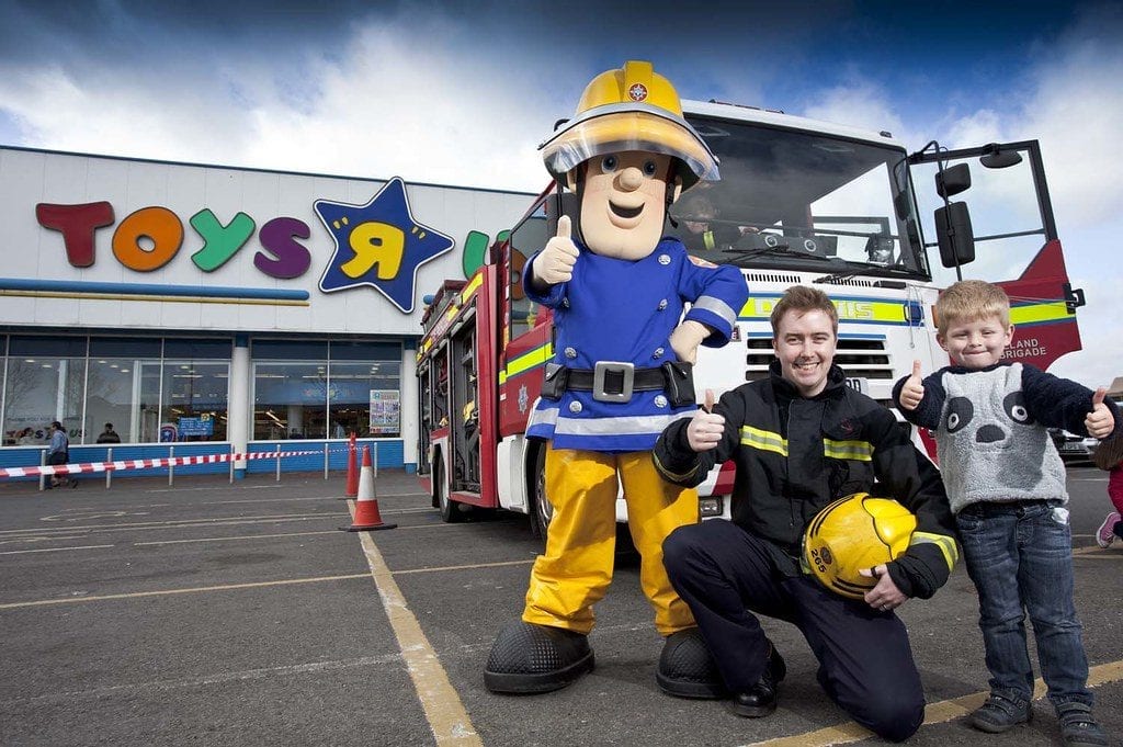 Toys: A person in a Fireman Sam costume, a firefighter, and a child with thumbs up in front of a fire engine and a 'Toys R Us' store.