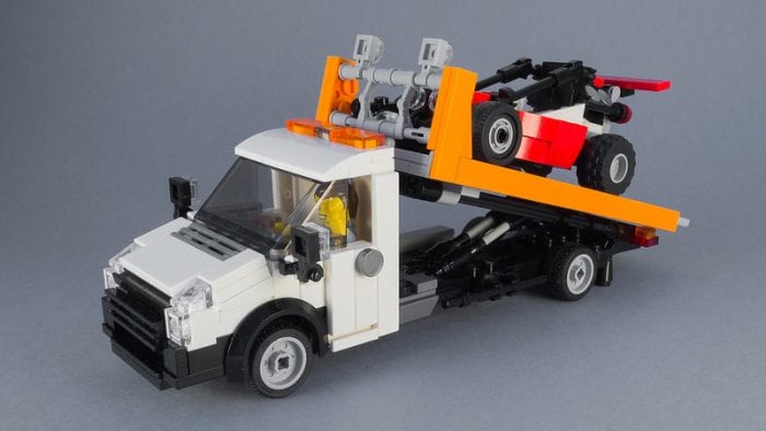 Toy Tow Truck: White and orange toy flatbed tow truck made from building blocks with a black and red car on top, set against a gray background.