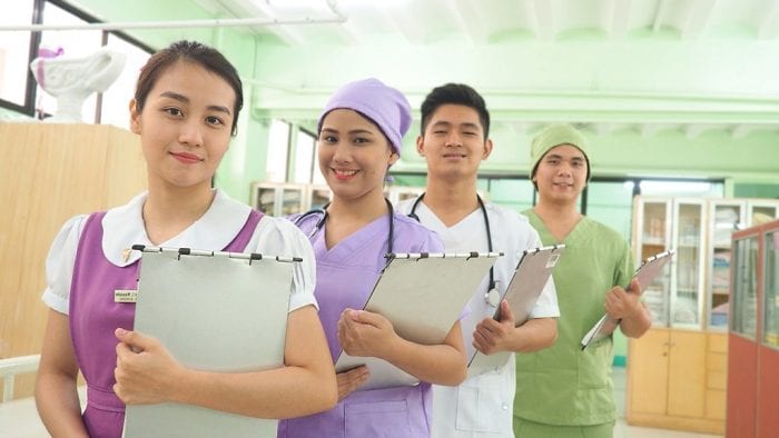 A group of four medical students in scrubs, holding clipboards and smiling confidently, standing in a hospital corridor.