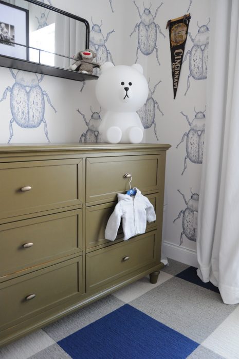 A chocolate brown dresser. There is a baby jacket hanging on the handle of the dresser. Above the dresser is a cute teddy bear. 