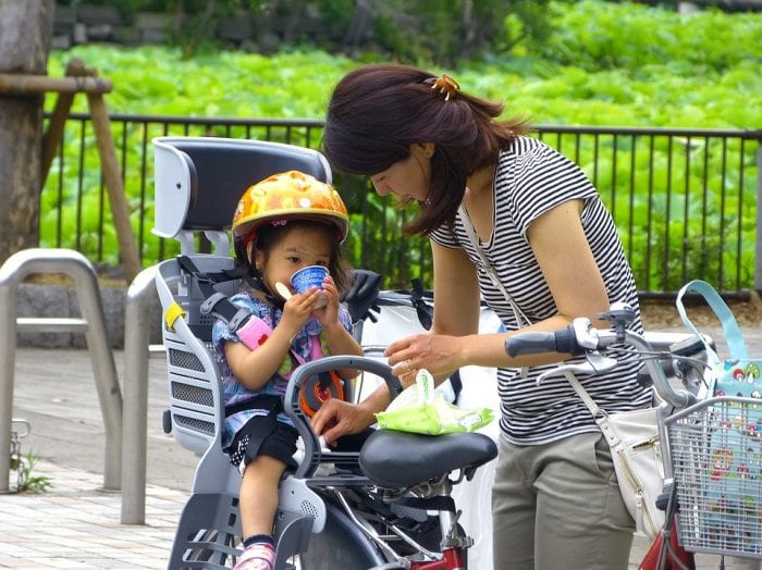 A mother feeding her daughter who is wearing a helmet while in a baby bike seat at the park. 