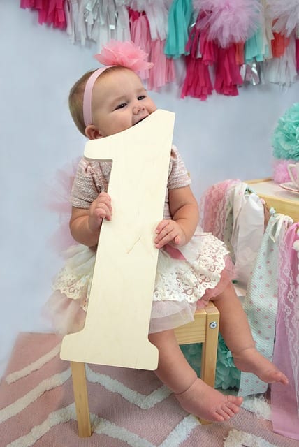 This is a photoshoot of a cute and adorable 1-year-old kid celebrating her birthday. She is holding a big number 1. She seems so happy.