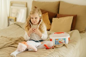 A kid pretends to be a doctor with her doll and other game sets on the bed. The kid looks happy while wearing the stethoscope. The color of the bed is brown. The bed looks soft and comfortable. 