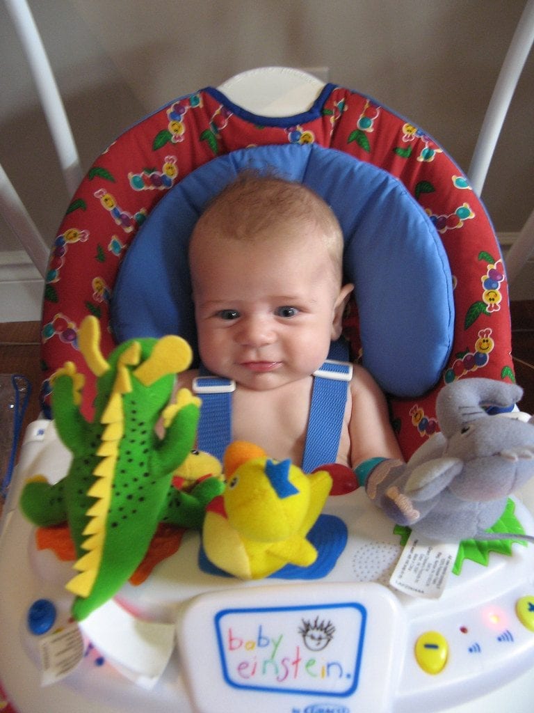 A little baby boy is looking at his dinosaur toy together with the yellow duck and elephant toy while setting on his rocker with red and blue color combination. 